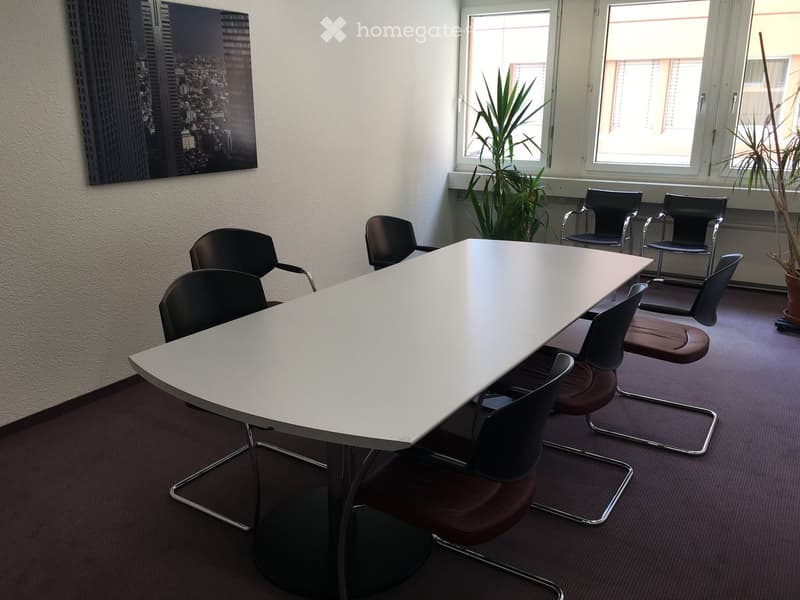 Conference room with a large white table surrounded by black chairs, large windows with plants, and a cityscape picture on the wall.