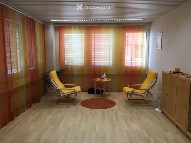 Bright living room with wooden flooring, two yellow chairs, small round table, fan, cabinet, and plant near the window.