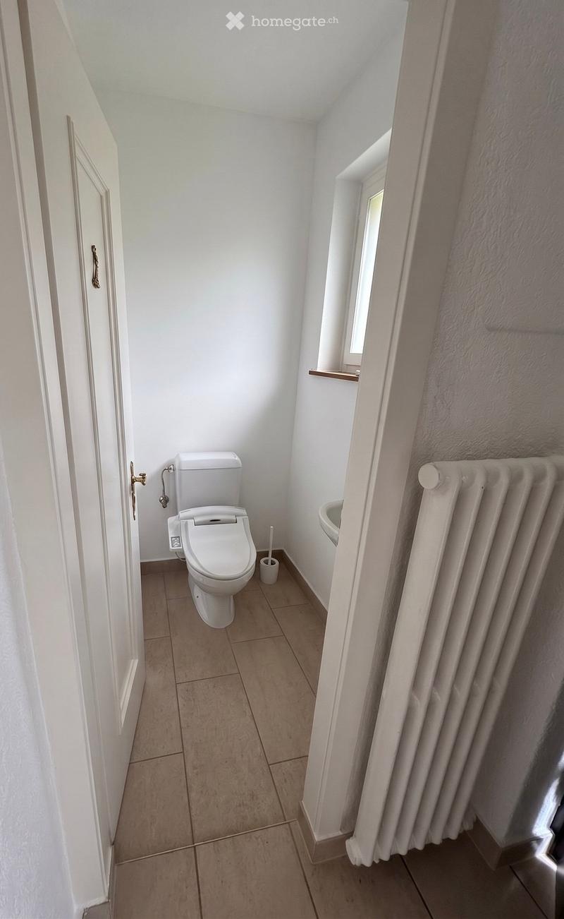 Modern bathroom with white tiled walls, beige tiled floor, toilet, sink, and radiator. Features a window and a door with a decorative handle.