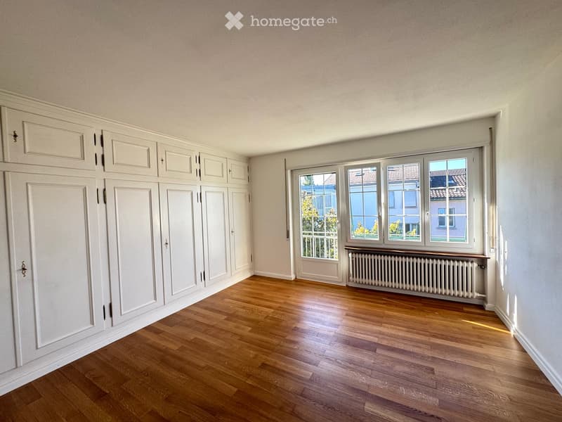 Empty room with wooden flooring, white built-in wardrobes, large windows with radiator below, and neutral-colored walls.