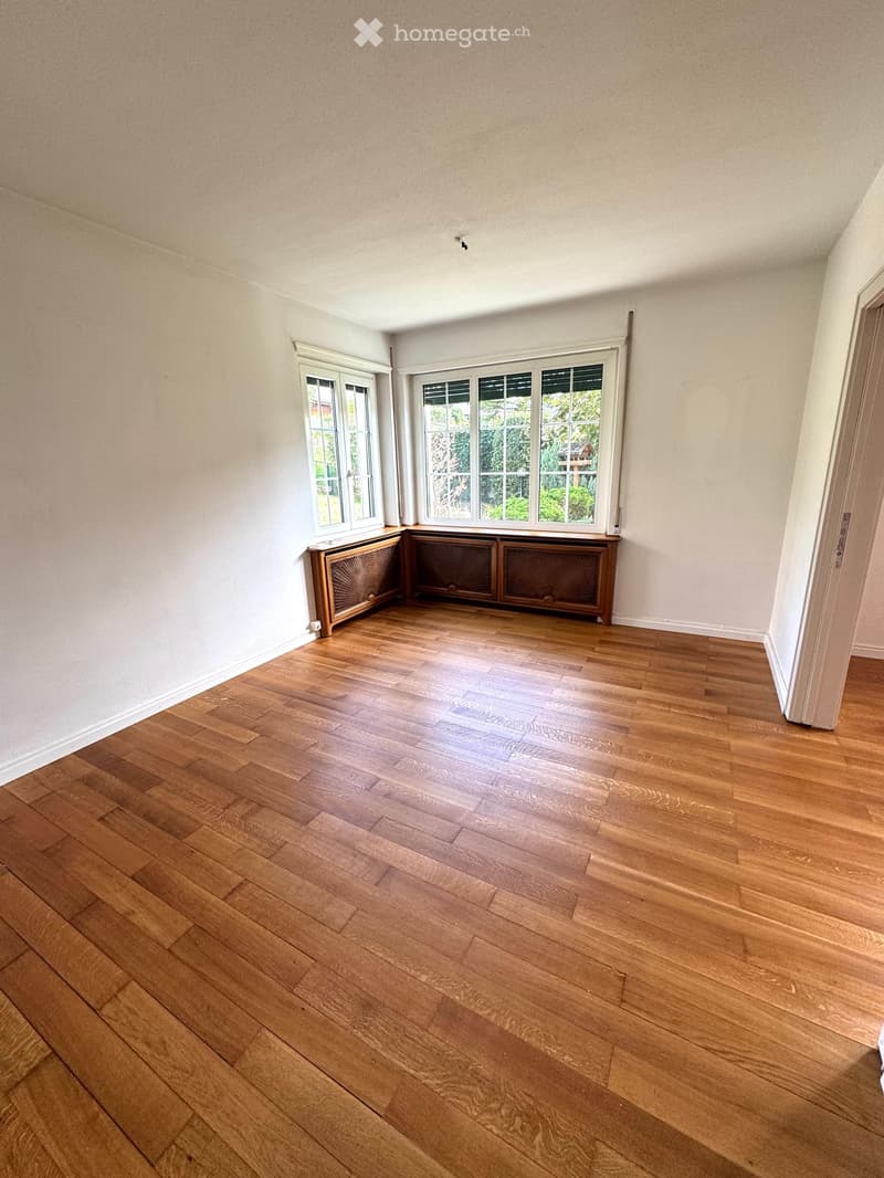 Empty room with wooden flooring, white walls, and large windows with radiators beneath them.