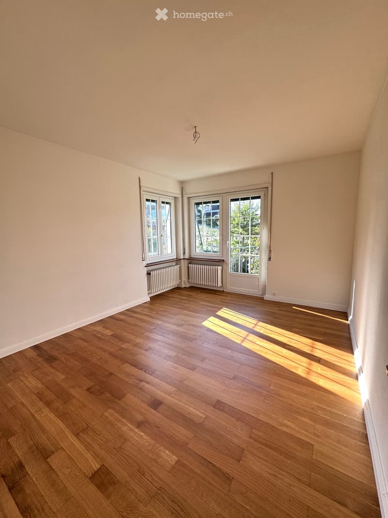 Empty room with wooden flooring, white walls, and large windows allowing natural light to enter. Radiators are visible under the windows.