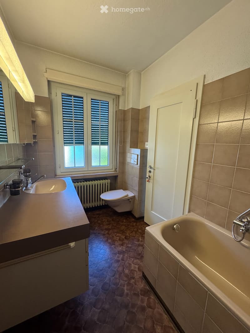 Bathroom with a sink, toilet, bathtub, mirror, and radiator. The room features tiled walls and a window with blinds.