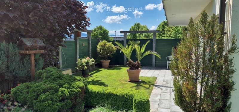 Well-maintained garden with a small lawn, potted plants, shrubs, and a wooden pergola. The area is enclosed by a green fence and features a pathway leading to a seating area.