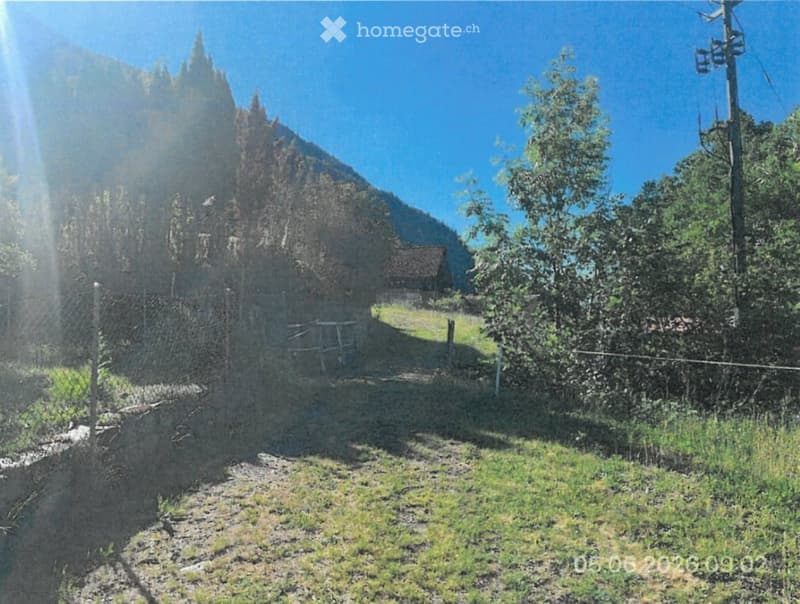 A rural outdoor area with a fenced yard, a small wooden structure, and a mountainous backdrop under a clear blue sky.