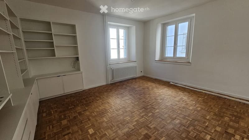 Empty room with wooden herringbone flooring, white walls, built-in shelving unit, and two windows with radiators below them.