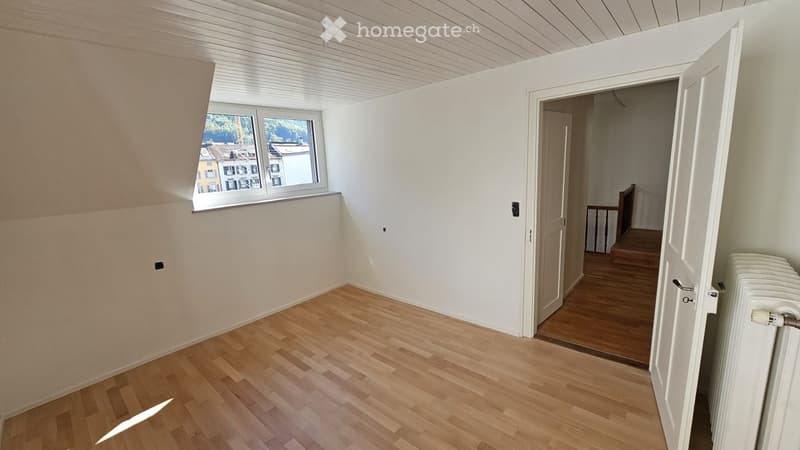 Empty room with wooden flooring, white walls, and a window offering a view of buildings outside. A door leads to another room with wooden flooring and a visible staircase.