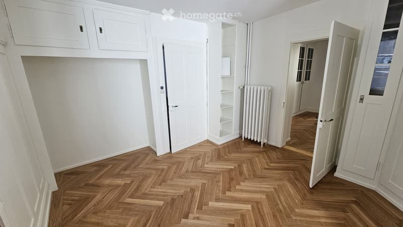 Empty room with herringbone wooden flooring, white walls, built-in cabinets, and a radiator. No furniture or personal items are present.