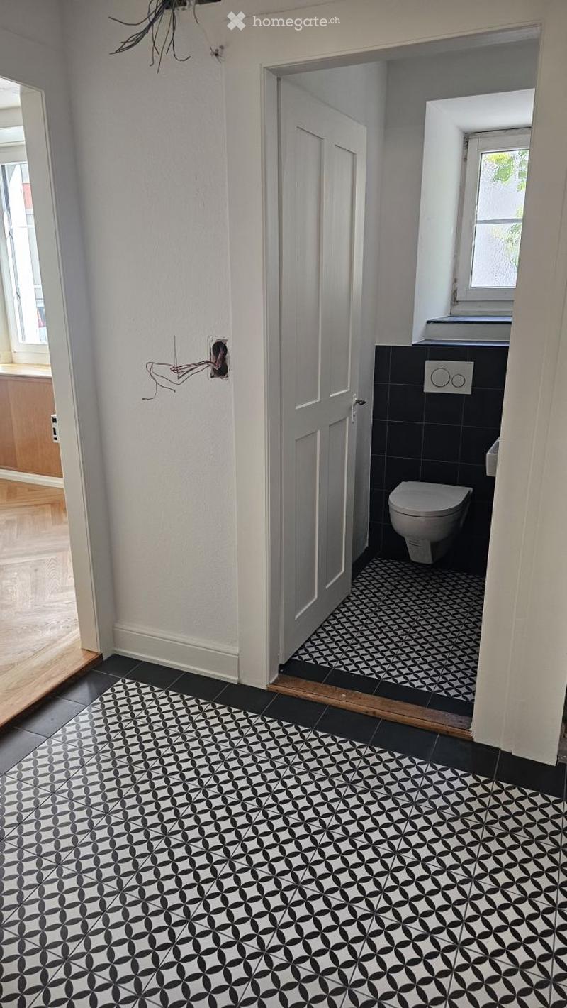 Hallway with black and white patterned tiles, leading to a bathroom with a toilet and black tiled walls.