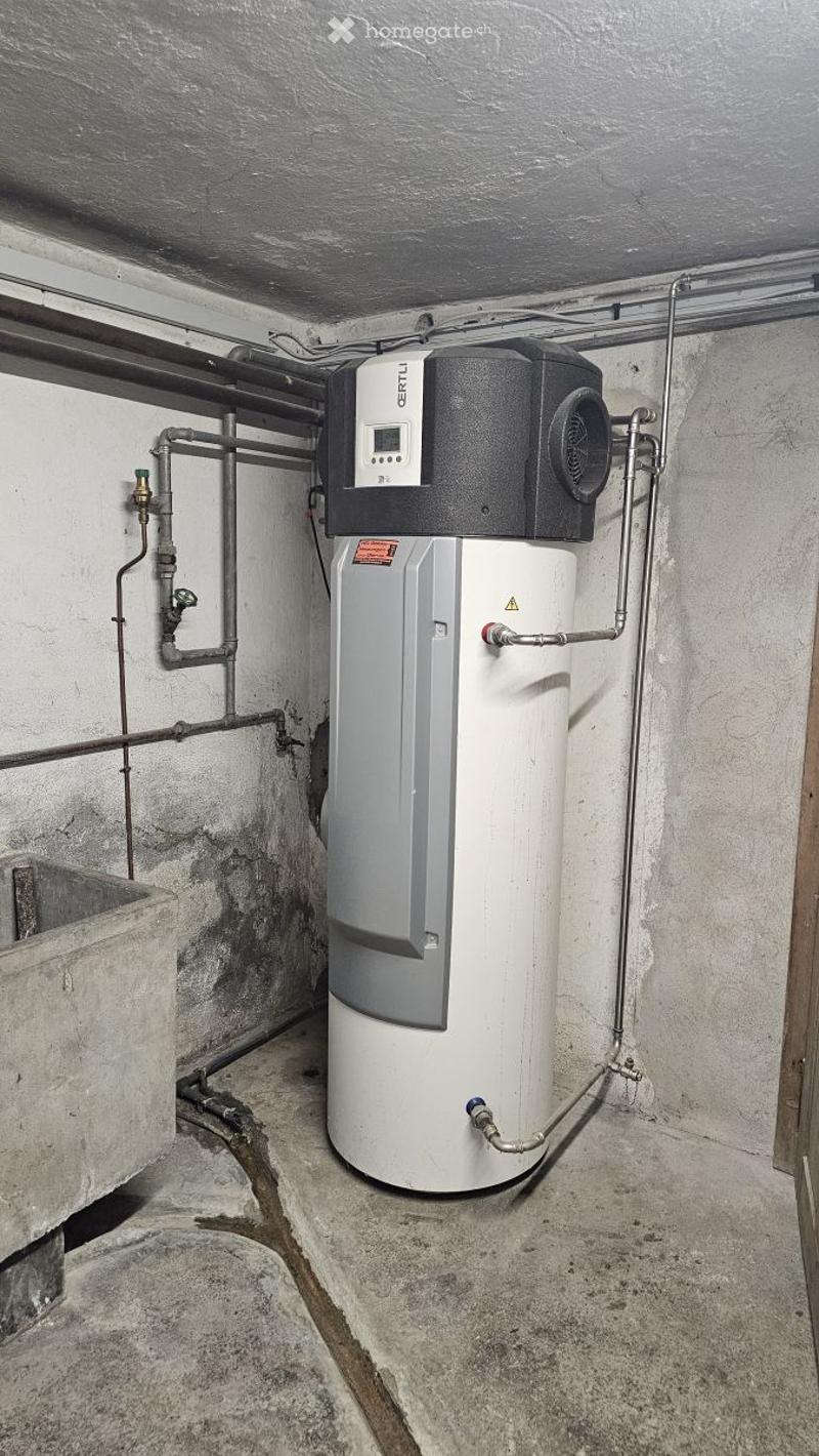 A modern water heater with a control unit mounted on top, surrounded by pipes and valves in a utility space.