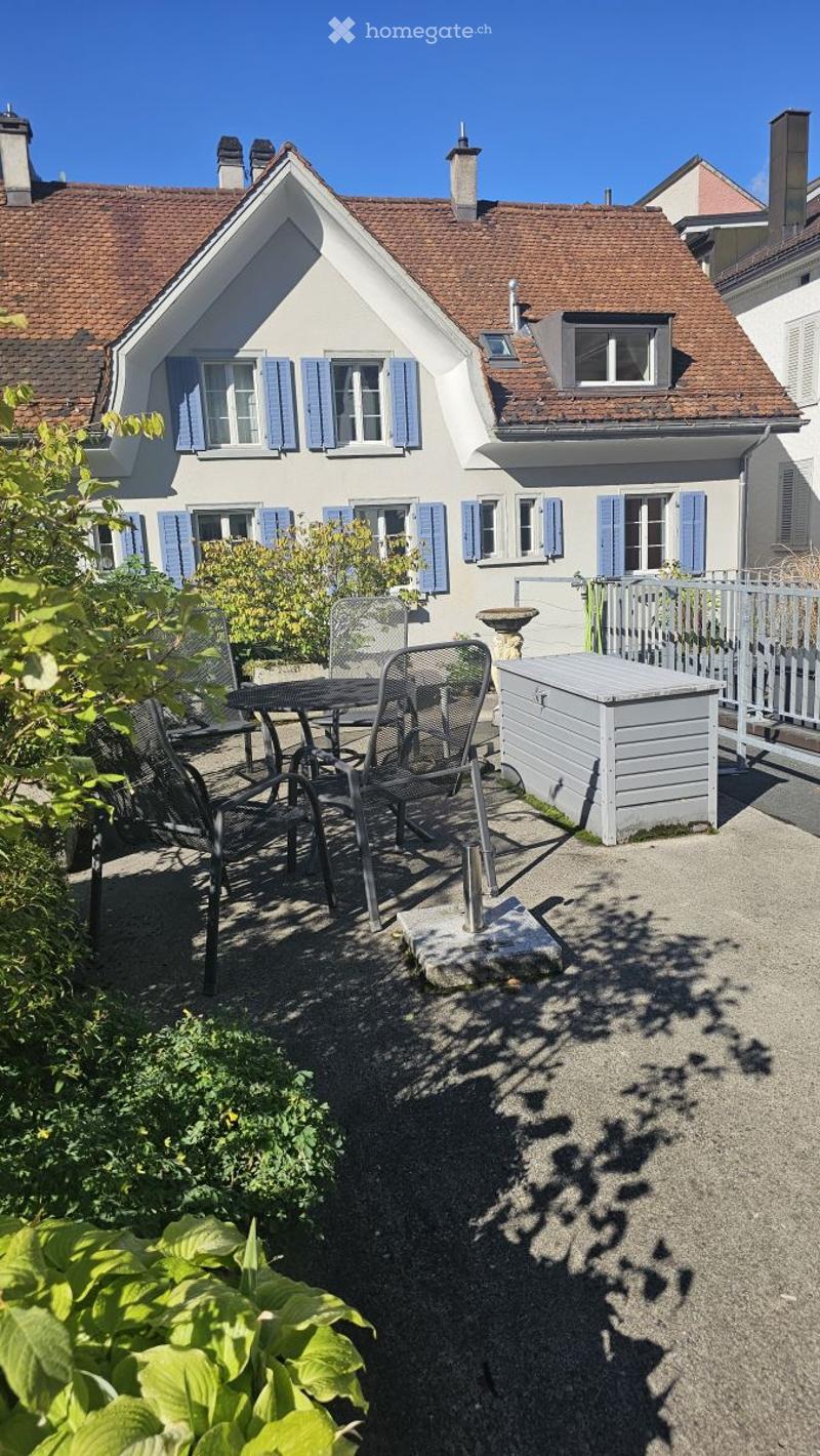 Outdoor patio area with a small table, chairs, and a storage box, surrounded by greenery and a white fence.
