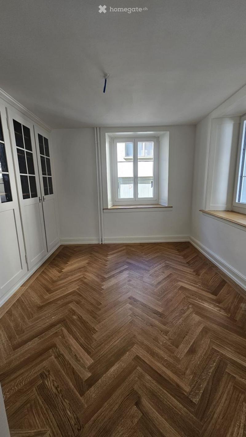 Empty room with herringbone wooden flooring, white walls, and large windows with sills.
