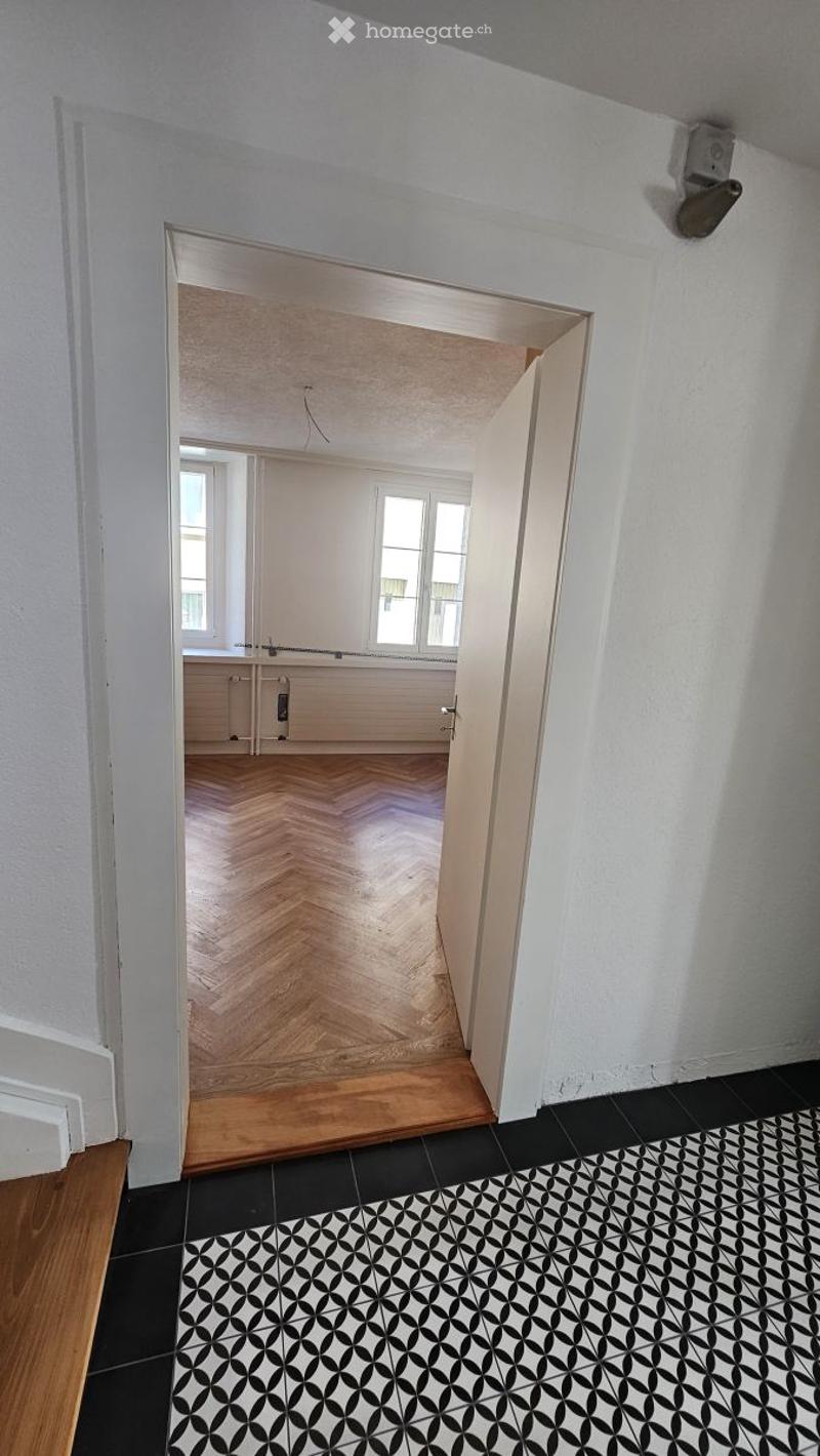 Empty room with white walls, wooden herringbone flooring, and a black and white patterned rug. The adjacent room is also unfurnished, with wooden flooring and white walls.
