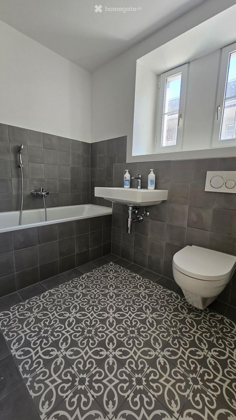 Modern bathroom with dark grey tiles, a white bathtub, a toilet, a sink with a faucet, and a decorative black and white patterned floor.