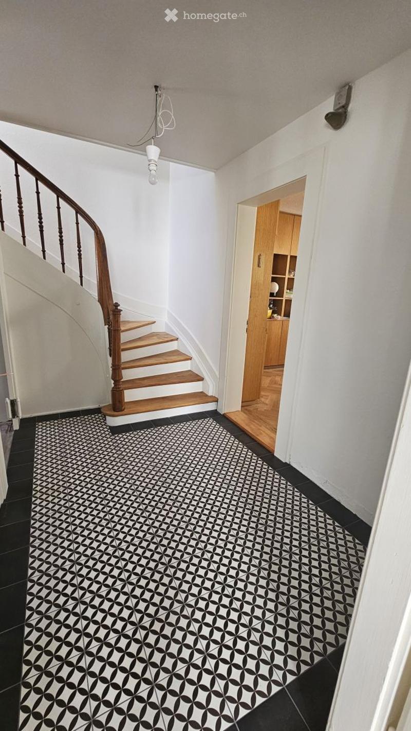 Hallway with black and white patterned tile floor, wooden staircase with wooden handrail, white walls, and a small open closet with wooden shelves.