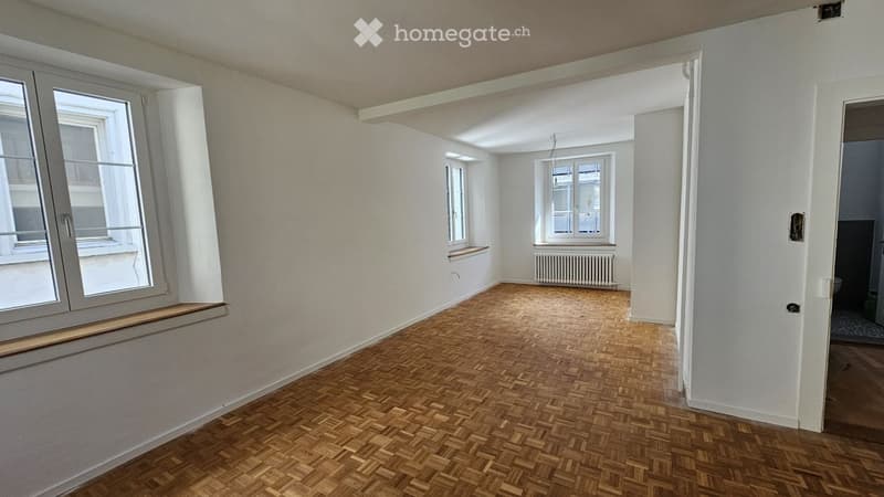 Empty room with wooden herringbone flooring, white walls, large windows, and a radiator. No furniture or equipment present.