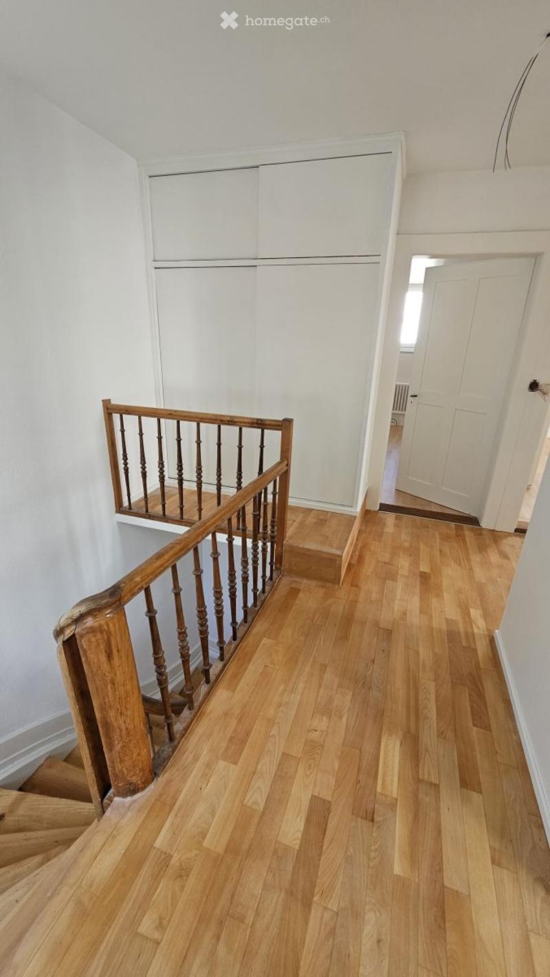 A well-lit hallway with wooden flooring and a staircase featuring a wooden handrail and balusters. The walls are painted white, and there is a closed white door at the end of the hallway.