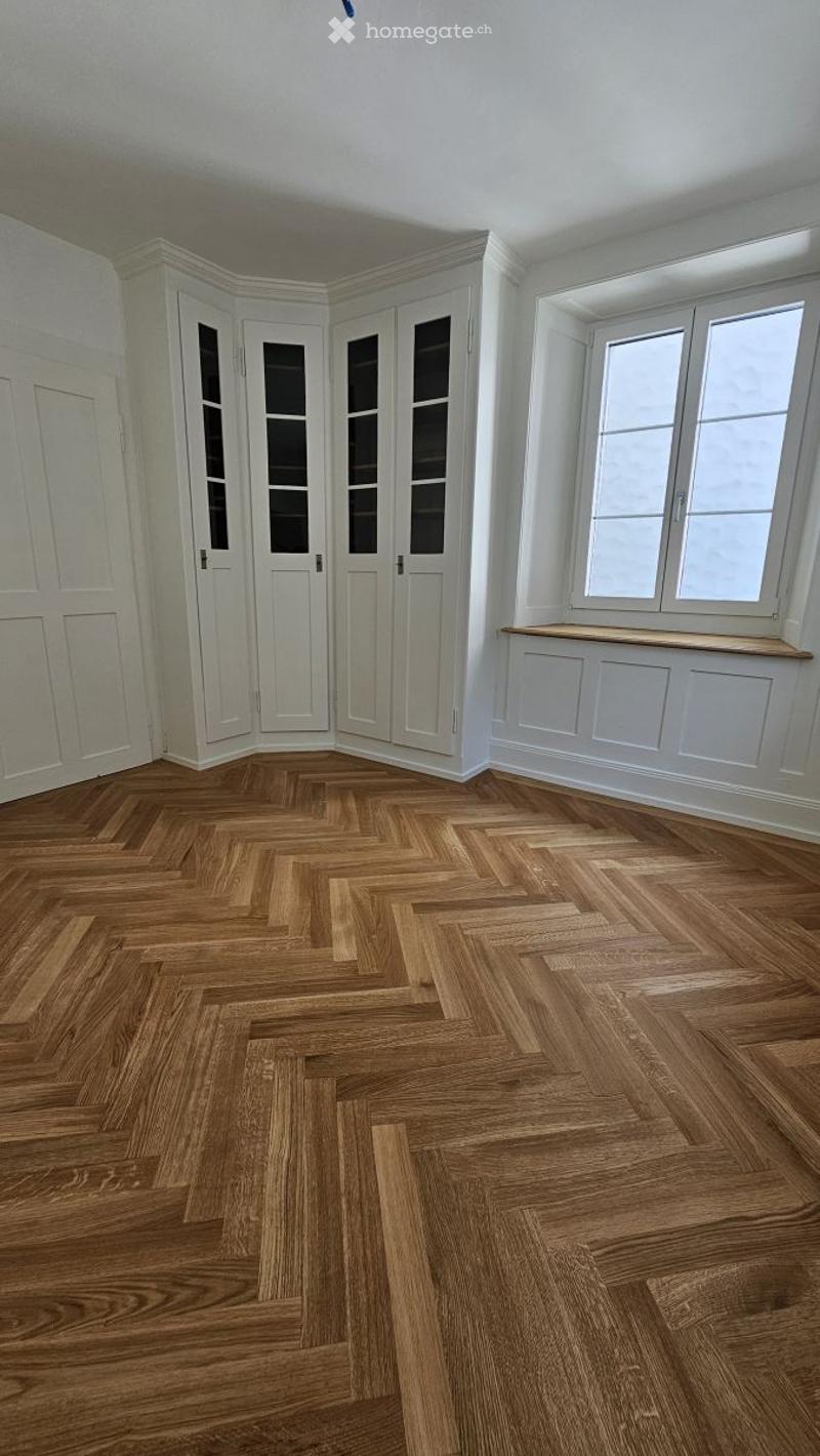 Empty room with herringbone wooden flooring, white walls, built-in white cabinets, and large windows with a window seat.