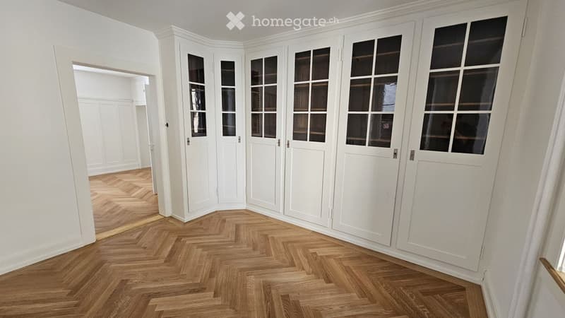 Empty room with white walls, herringbone wooden flooring, and built-in white cabinetry with glass-paneled doors.