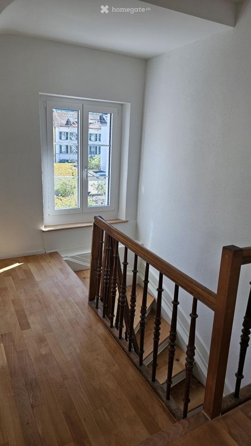 Empty hallway with wooden flooring, white walls, and a staircase with wooden railings leading upwards. A window at the end provides natural light.