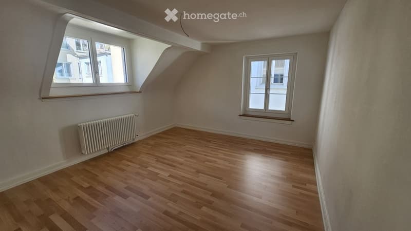 Empty attic room with wooden flooring, white walls, and two windows, one with a slanted roofline.