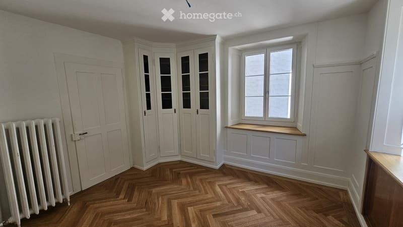 Empty room with wooden herringbone flooring, white walls, built-in corner cabinet, window with built-in bench, radiator, and door.