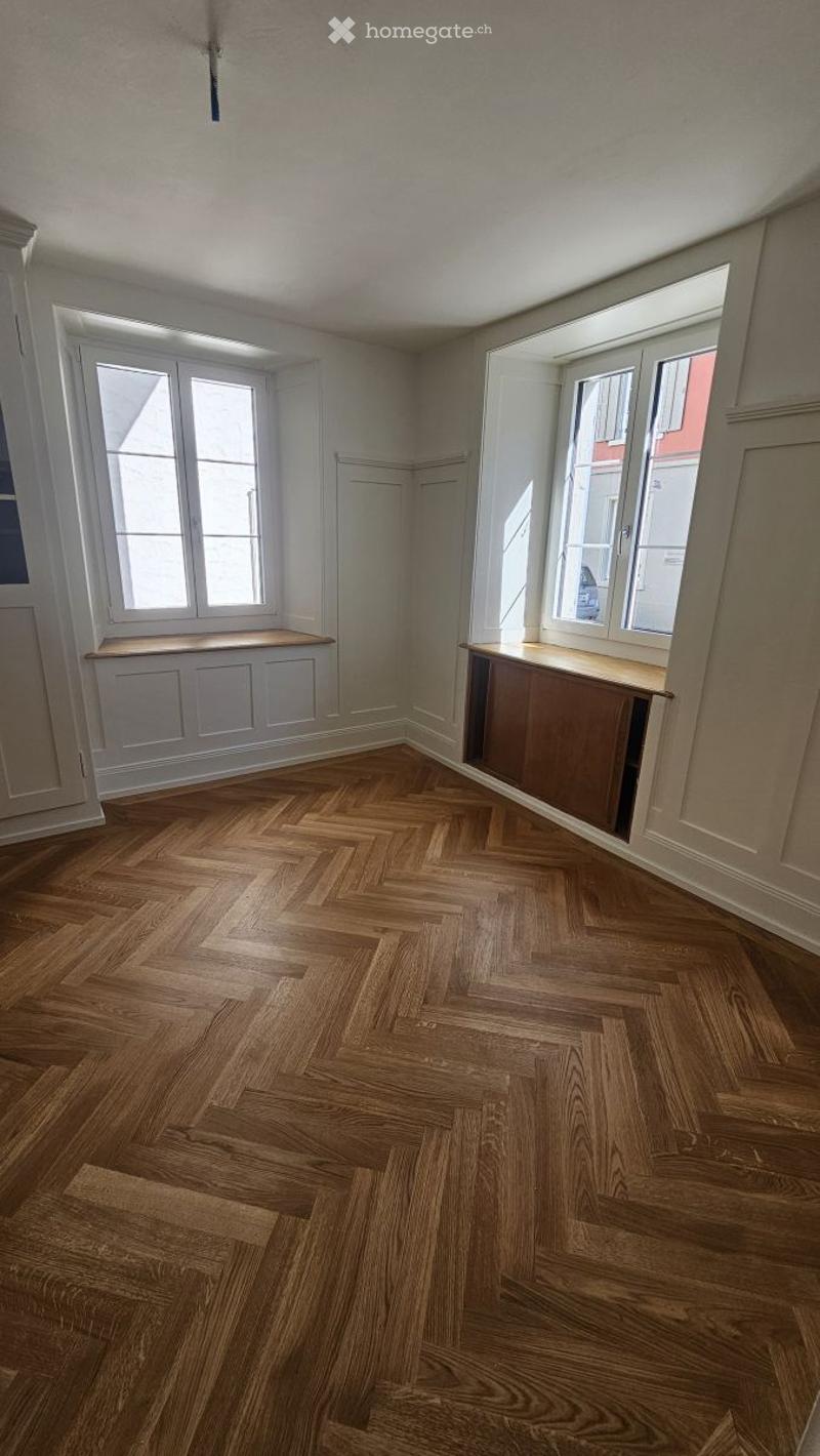 Empty room with herringbone wooden flooring, white walls, and large windows with built-in benches.