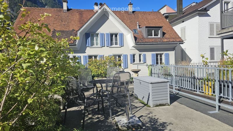 A well-maintained garden area with a patio, outdoor furniture, and a view of a charming house with blue shutters and a red-tiled roof.