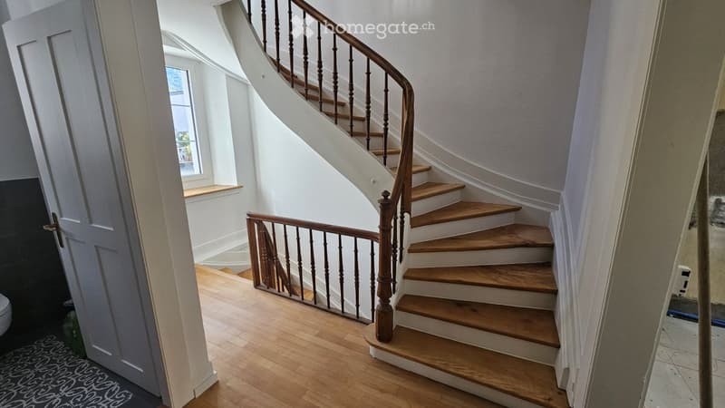 Spacious hallway with wooden staircase featuring a polished wooden handrail and balusters, leading to an upper level. The hallway has light-colored walls and wooden flooring.
