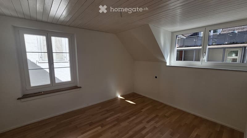 Empty room with wooden flooring, white walls, and two windows allowing natural light. The ceiling is slanted, typical of an attic or loft space.