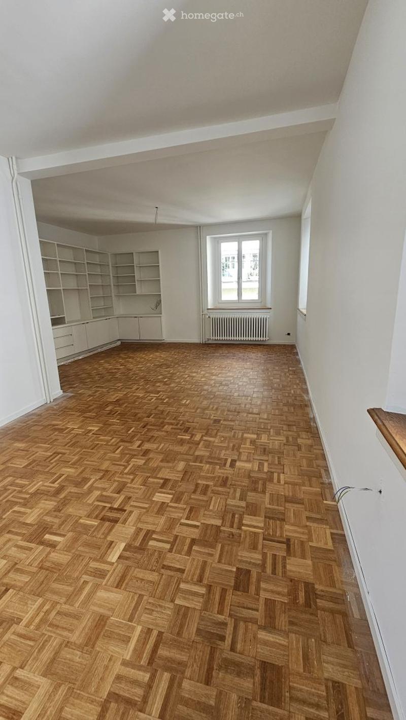 Empty room with wooden herringbone flooring, white walls, built-in shelving unit, and a window with radiator below.