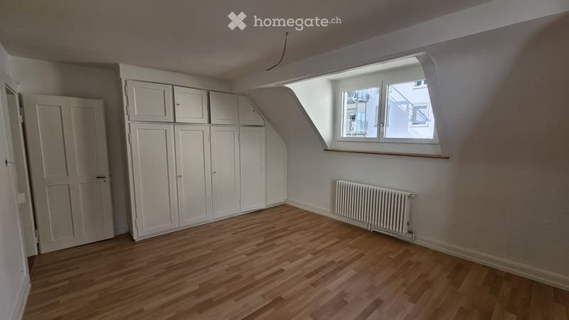 Empty attic room with wooden flooring, white built-in wardrobes, a window with a view, and a radiator.