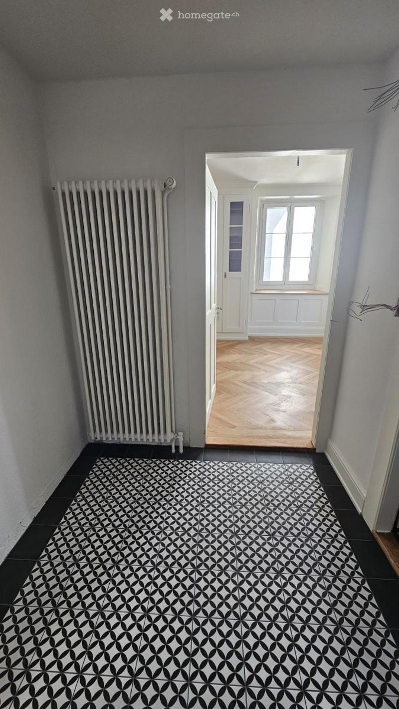 Empty hallway with black and white patterned tile floor, white radiator on left wall, and an open doorway leading to another room with wooden flooring and white walls.