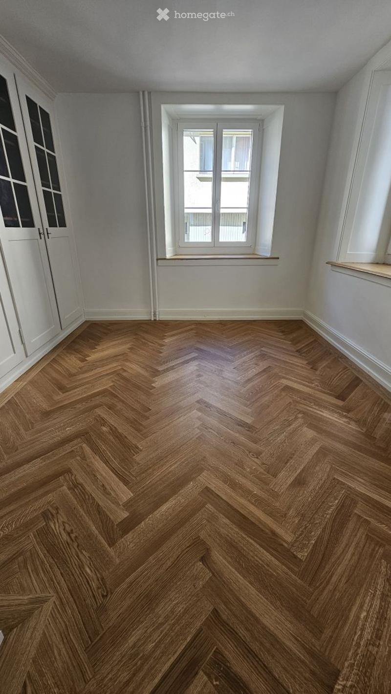 Completely empty room with herringbone wooden flooring, white walls, and large windows with white frames.