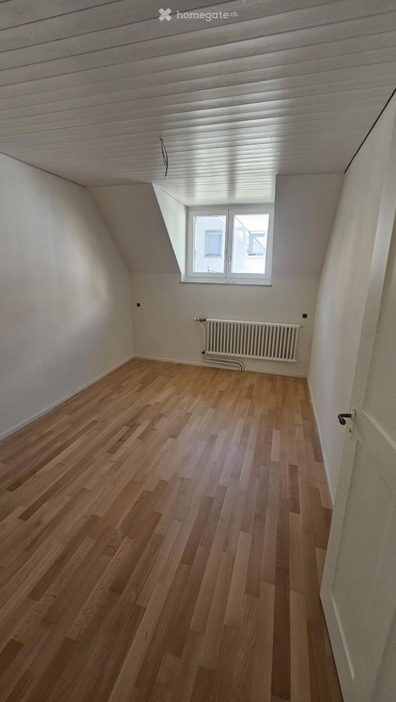 Empty attic room with wooden flooring, white walls, and a single window with white frame. No furniture or equipment present.