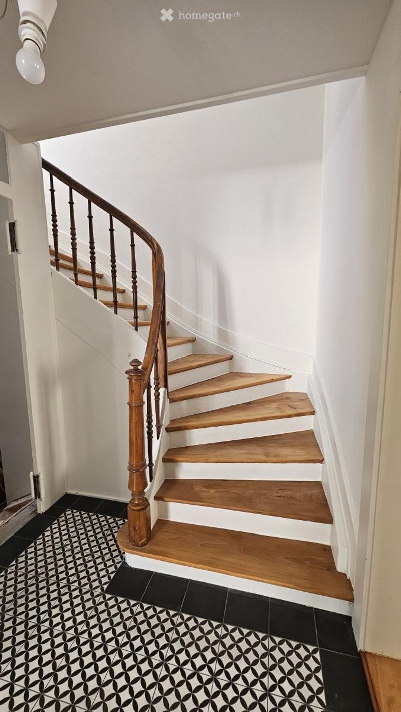 Wooden staircase with a dark wooden handrail and balusters, white walls, black and white patterned tile floor, and a light fixture on the ceiling.