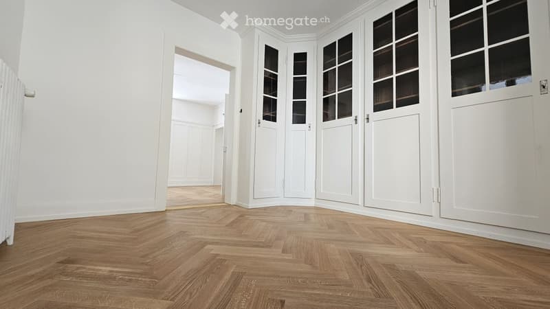 Completely empty room with white walls, wooden herringbone flooring, and white cabinetry with glass doors.