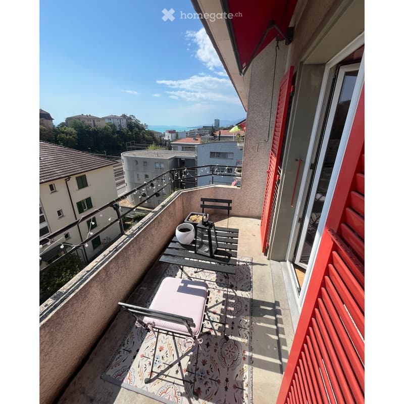 Balcony with a table, chair, pot, and bench, overlooking a cityscape with buildings and greenery.