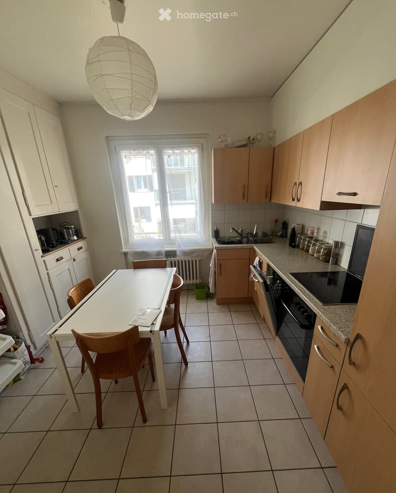 A modern kitchen with light wooden cabinets, a white dining table with four chairs, and various appliances including an oven, dishwasher, and sink.