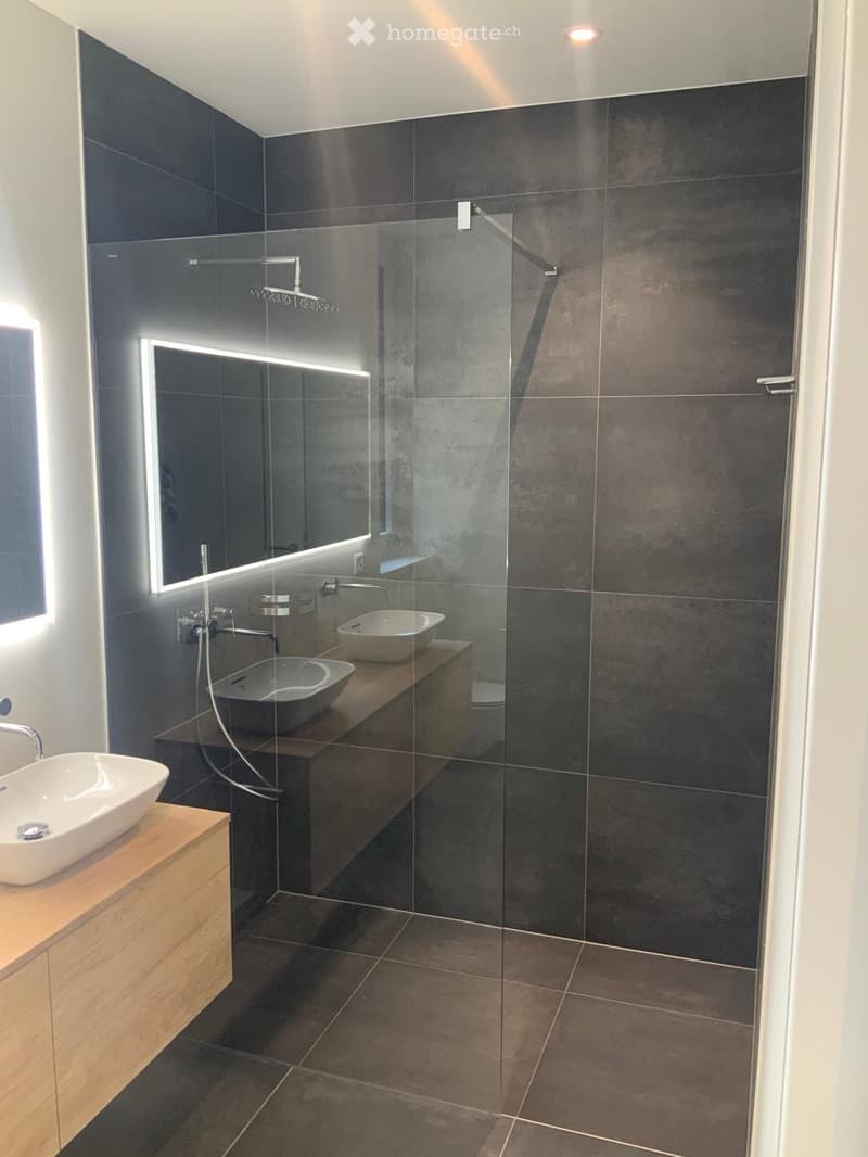 Modern bathroom with dark grey tiles, a glass-enclosed shower, two sinks on wooden vanities, and a large illuminated mirror.