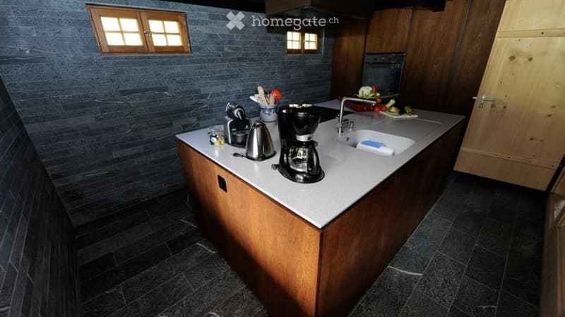Modern kitchen with dark grey tiles, white countertops, wooden cabinets, and various appliances including a coffee maker, sink, and oven.