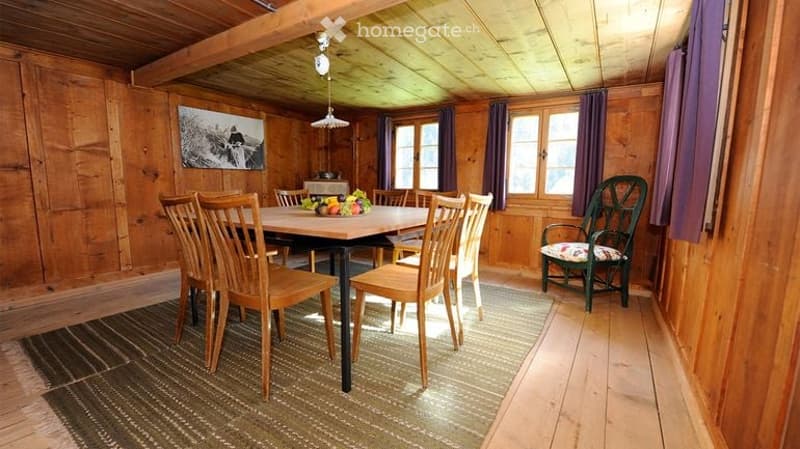 Cozy wooden-paneled room with a dining table surrounded by chairs, a single armchair, and a fruit bowl on the table. The room features wooden walls, a wooden ceiling with exposed beams, and large windows with purple curtains.