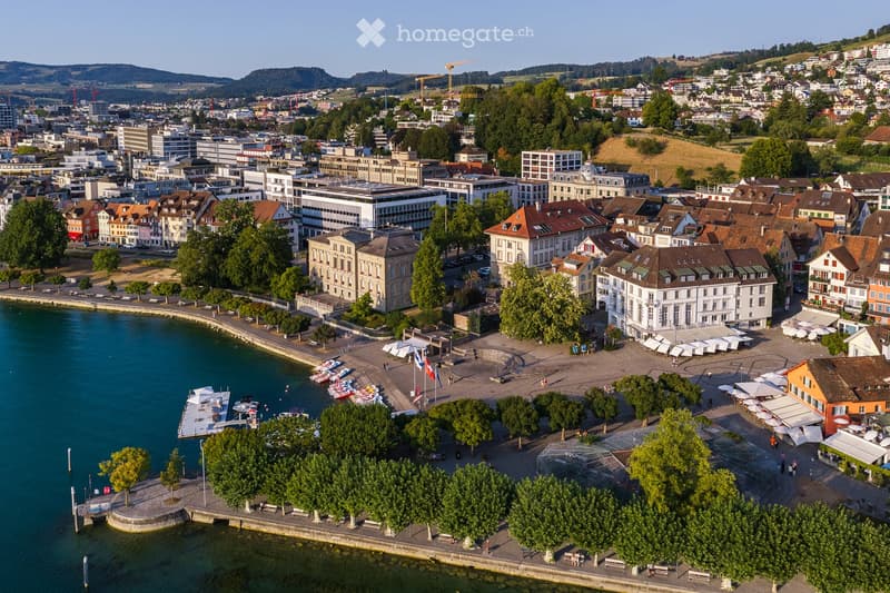 Aerial view of a cityscape with a waterfront, featuring a mix of residential and commercial buildings, trees, boats, and a promenade area.