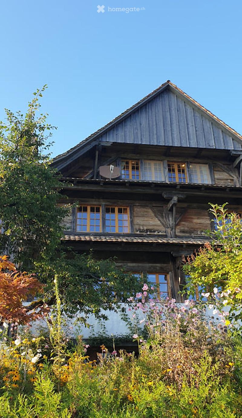 A two-story wooden house with a steeply pitched roof, large windows, and a balcony on the upper floor, surrounded by lush greenery and flowers.