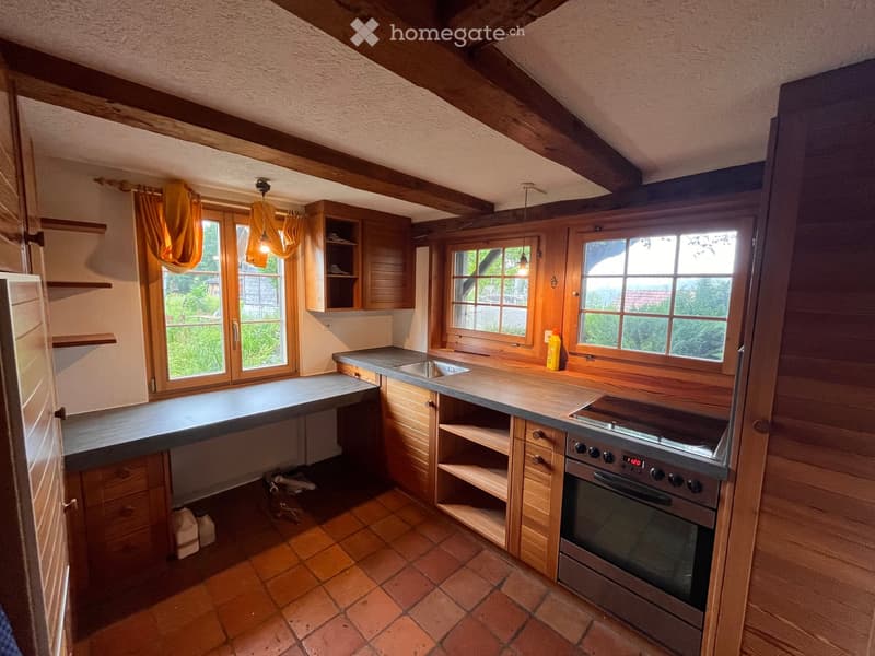 Cozy kitchen with wooden beams, exposed wooden cabinets, a tiled floor, and a modern oven. The space features large windows with orange curtains and a countertop with a sink.