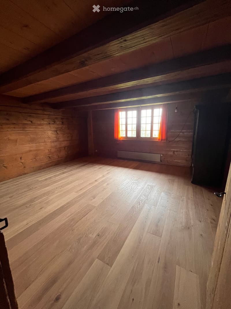 Empty attic room with wooden walls and floor, large windows with orange curtains, exposed wooden beams on the ceiling, and a radiator under the windows.