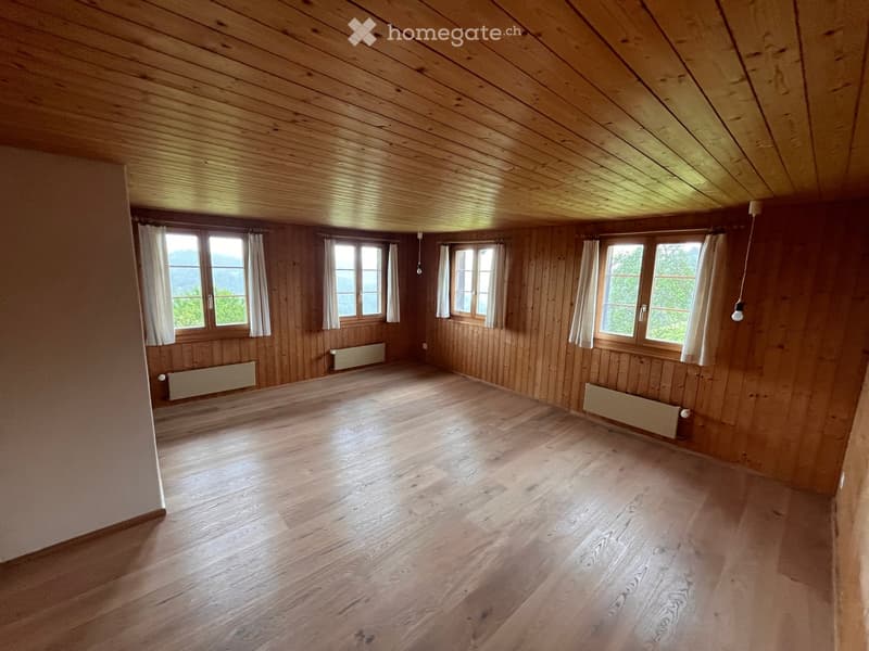 Empty attic room with wooden walls, wooden floor, and large windows with white curtains. Natural light illuminates the space.