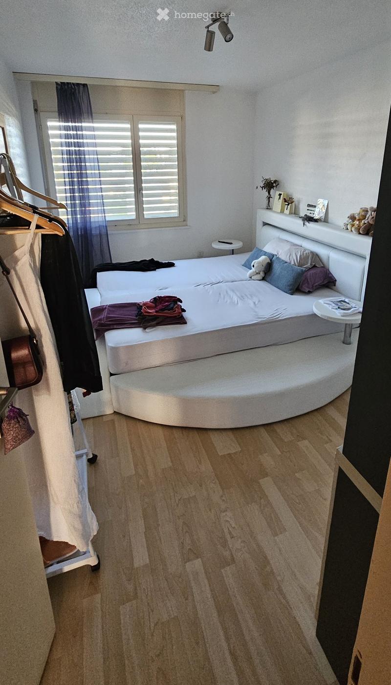 Modern bedroom with a round bed, nightstand, small table, and wardrobe. Wooden flooring and large window with blinds.