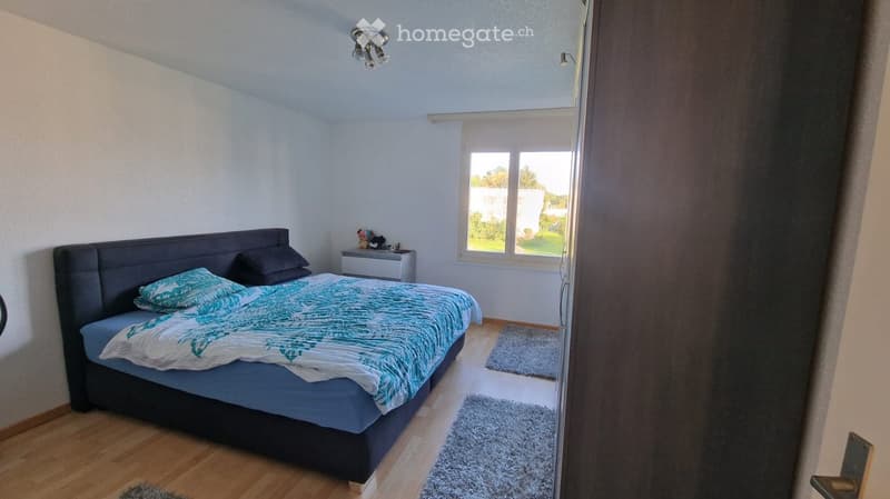Bedroom with a large bed, nightstand, and two rugs on the floor. The room has light-colored walls and wooden flooring.