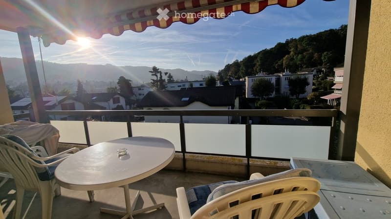 Balcony with a table and chairs, overlooking a scenic town and hills under a clear sky.